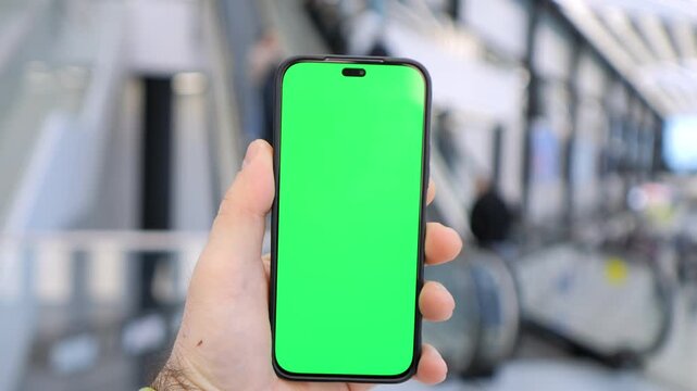 Hand holds phone with green screen. Background shows blurred train station. Three keyframes show slight camera movement. People walk past in background. No animals or plants visible