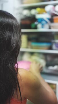 Unrecognizable woman with dark hair opening a refrigerator to get a meal from a food container