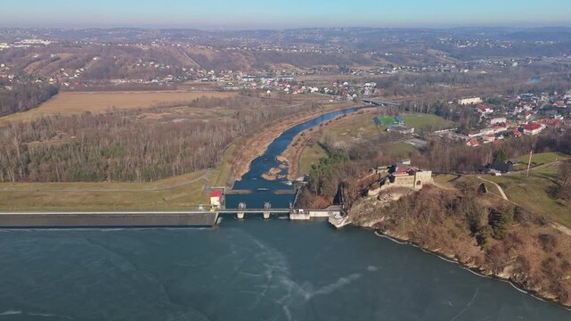 At Lake Dobczyce in Poland, the reservoir and its dam sit beside the Raba River with Dobczyce Castle overlooking surrounding winter hills and forests, seen in a drone view.