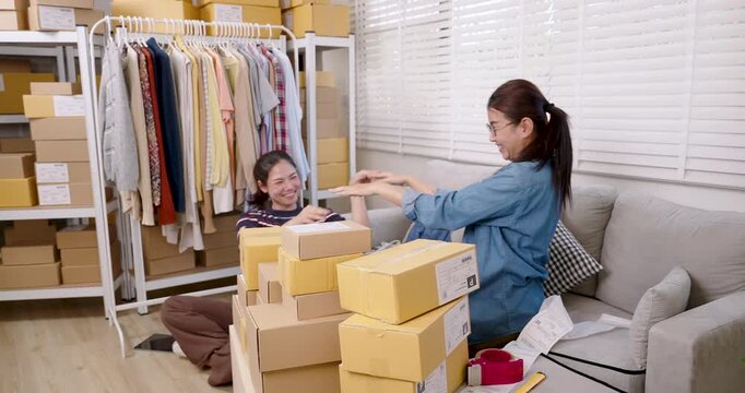 Asian women online business partners smile during parcel box preparation, sharing positive moment after completing orders, supporting strong teamwork inside home based e-commerce workflow