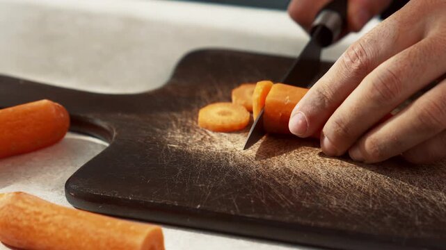Close-up footage of a person's hands slicing a fresh carrot with a knife on a rustic wooden cutting board.