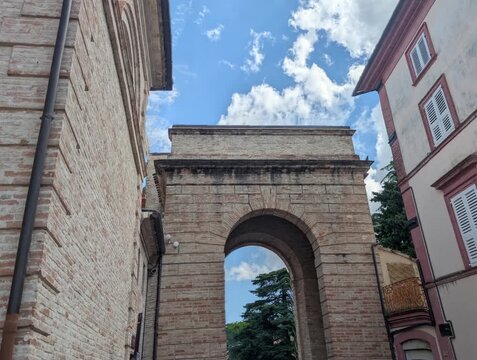 L&rsquo;ingresso storico di Urbisaglia si apre con l&rsquo;arco in mattoni e la strada che va verso il centro. Una scena luminosa che unisce architettura antica e vita quotidiana marchigiana.