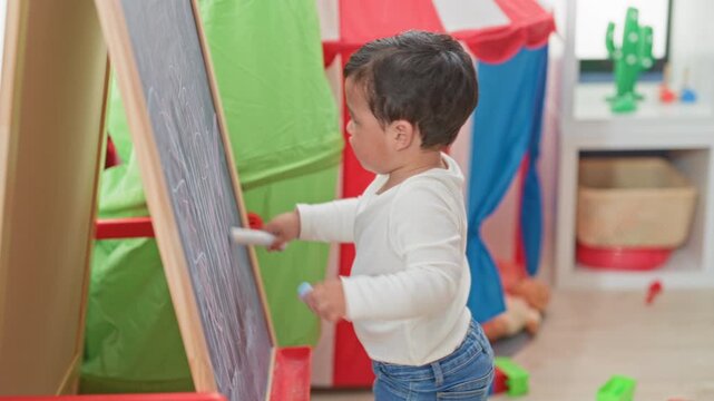 Boy drawing on chalkboard holding chalk and scribbling at an easel in a colorful playroom inside a building; focus learning curiosity.