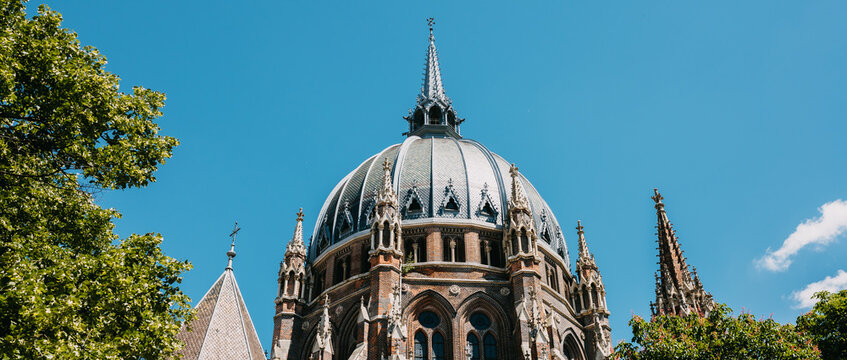 Historic cathedral dome rises above leafy trees under blue sky, revealing ornate brickwork and view dome