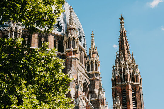 Historic brick cathedral rises with ornate towers, pointed spires, and leafy trees against bright blue