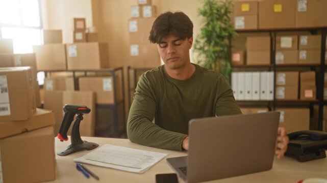 Man at desk checking parcels and typing on laptop while handling shipping paperwork with barcode scanner and stacked boxes in building; entrepreneurial focus.