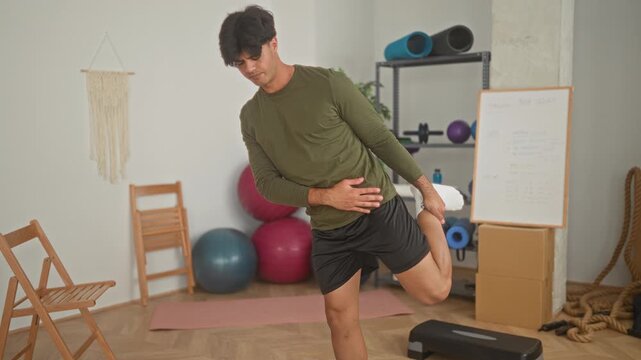 Man holds foot to glute with hand while balancing on one leg in a home studio with yoga mat, exercise balls, stepper and whiteboard; focus balance.