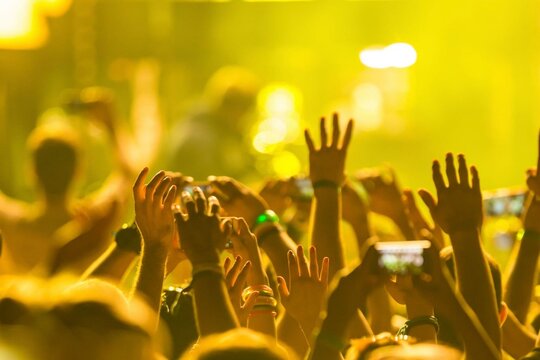 Golden lights illuminate a crowd's raised hands. Energetic faces glow with excitement and joy. People capture the moment on phones and flashlights. Music pulses through the air, connecting all