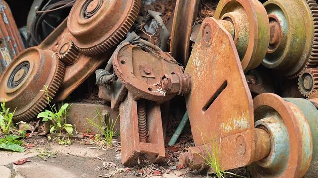 A collection of rusty metal parts is piled together in a disused area filled with greenery. The discarded items reveal a history of industrial use, with a focus on their weathered textures and forms
