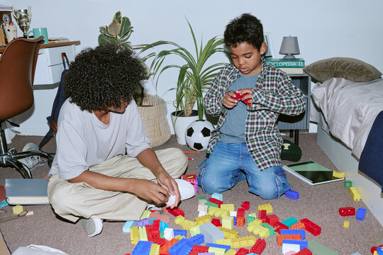 Wide shot of African American teenage boy and younger brother playing with building blocks on bedroom floor, supporting learning. Ideal for parenting, child development, home education marketing