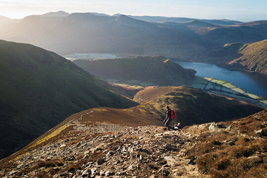 A female hiker and their dog walking down from the summit of Grasmoor with Whiteless Pike, Rannerdale and crummock Water in the distance on a bright winters morning in the Lake District, UK