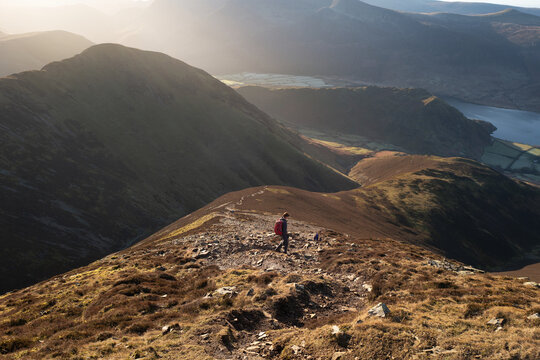 A female hiker and their dog walking down from the summit of Grasmoor with Whiteless Pike and Rannerdale  in the distance on a bright winters morning in the Lake District, UK