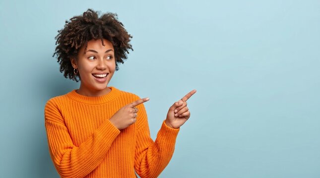 Excited young woman with afro hair wearing orange sweater pointing fingers to copy space on blue background, look there