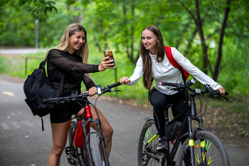 Fototapeta premium Two smiling female friends taking a selfie with a smartphone while standing on their mountain bikes in a lush green forest park.
