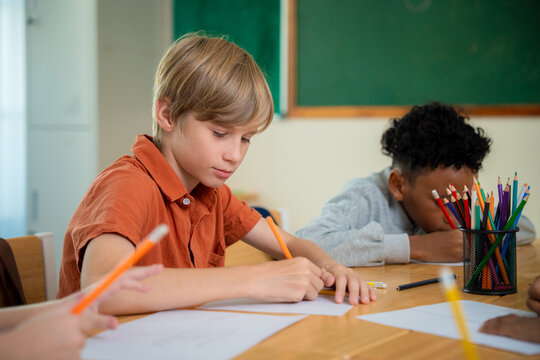Elementary school boy writing on paper during a lesson in classroom with classmates. Cute primary student concentrating on his exam or drawing at a desk with colorful pencils foreground.