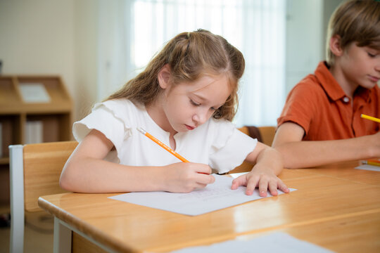 Elementary school girl concentrating on writing her homework or exam at a desk. Cute primary schoolgirl using a pencil to write on paper during an intensive lesson in classroom.