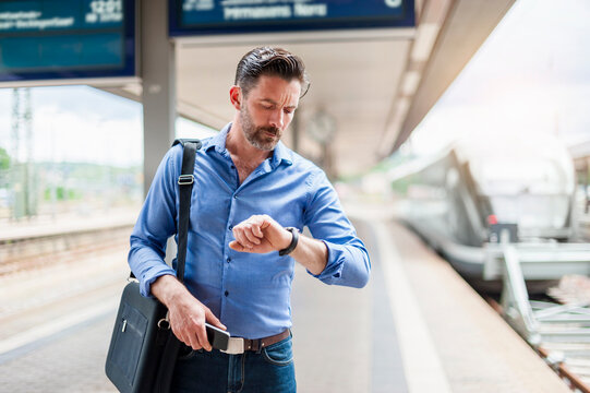 Bearded commuter looking at wristwatch while standing railroad station platform