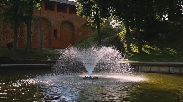 A water fountain sprays water high into the air, creating ripples on the surface of a pond. Surrounded by trees and a brick structure, this park offers a natural backdrop for relaxation