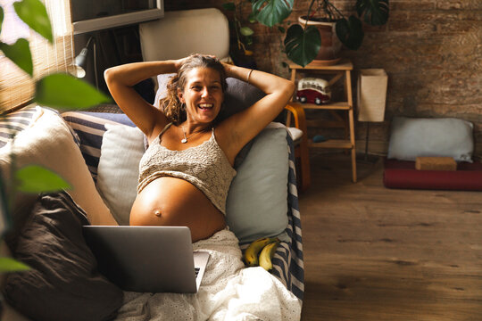 Smiling pregnant woman with laptop sitting on sofa at home
