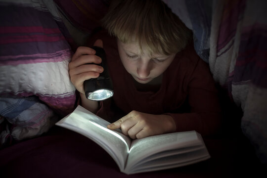 Boy with torch reading book under a blanket