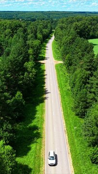 White car driving down a narrow asphalt road surrounded by lush green trees under blue sky