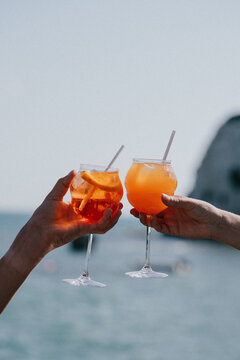 Two hands clinking glasses of orange spritzer drinks on a beach