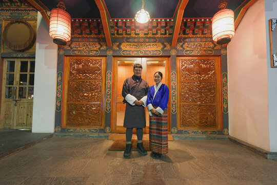 A happy young Asian couple wearing traditional Bhutanese clothing (Gho and Kira) smiling at the camera, standing together in front of an entrance decorated with intricate wooden carvings and colorful 