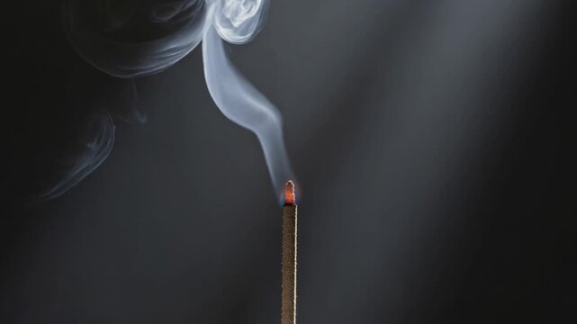 Incense stick burning with swirling smoke against dark background and light rays