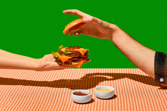 Hands assembling cheeseburger with sauces on checkered table. Concept of fast food, burger delivery, restaurant menu, meal preparation, and snack advertising.