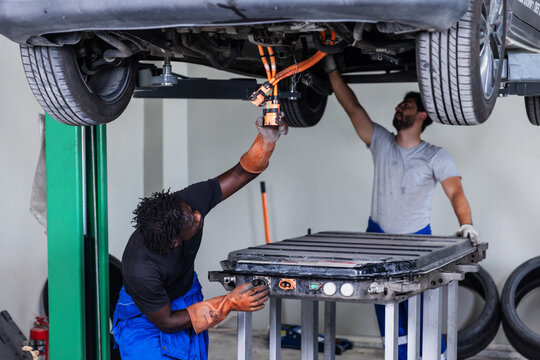 Professional EV technician disconnecting and inspecting a high-voltage battery connector beneath a lifted electric vehicle during maintenance.
