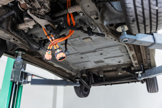 Close-up underside view of an electric vehicle lifted on a hydraulic service lift, revealing high-voltage orange cables and electrical connectors during maintenance inspection.