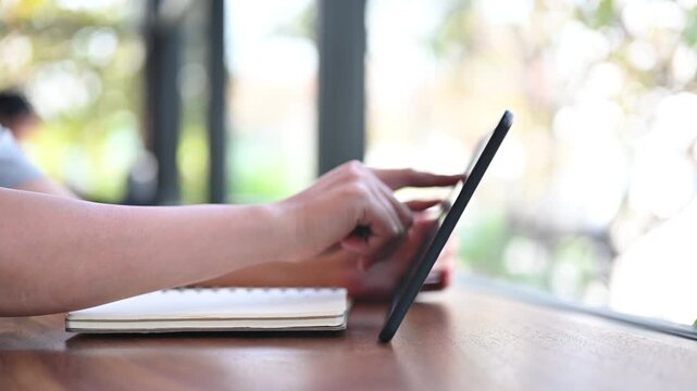 Person uses a digital tablet while sitting at a wooden table with a notebook nearby in a bright room.