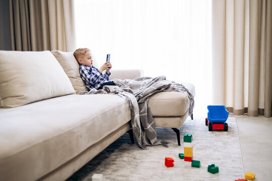 Young boy absorbed in smartphone on living room sofa, ignoring toys on floor&mdash;modern screen-focused childhood scene