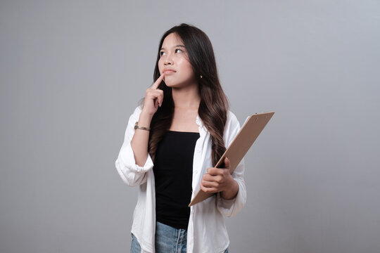 Thoughtful young Asian businesswoman holding clipboard and looking away on gray background