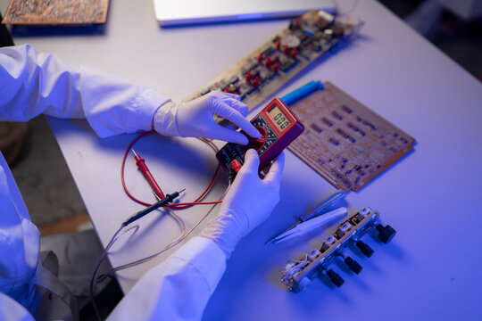 A person wears gloves and uses a multimeter to test a circuit board in a lab at night while other tools are nearby