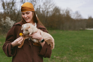 Fototapeta premium Joyful woman outdoors pets a gentle goat kid, holds a dandelion, sharing a warm moment of affection in a pastoral field, rustic jacket and scarf, serene countryside backdrop, candid lifestyle scene.
