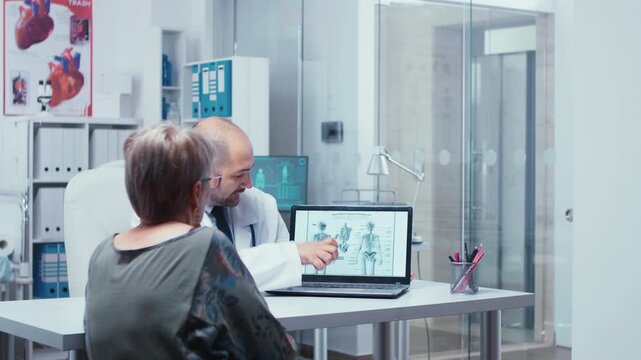 Doctor explains the human skeleton to an elderly patient using a laptop booklet while medical staff work in the background of a private clinic, with scan review and specialist medical advice.