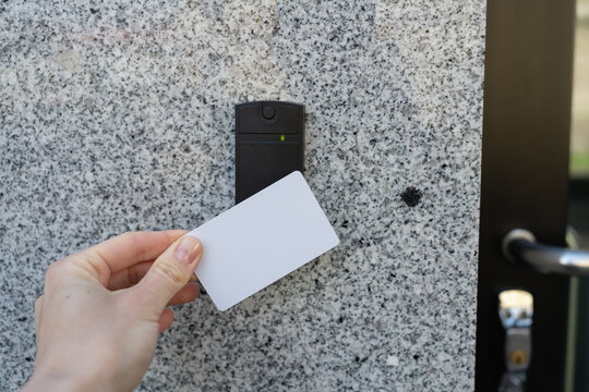 Closeup of a person's employee's hand holding a white access key pass card near a reader access device on a grey granite wall and a black glass door locked door, green light indicates granted access