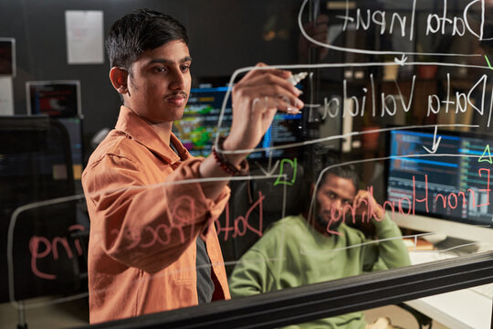 Asian young man writing data flow notes on glass board while colleague reviewing code on monitors, planning software project. For tech teamwork, agile development, startup marketing