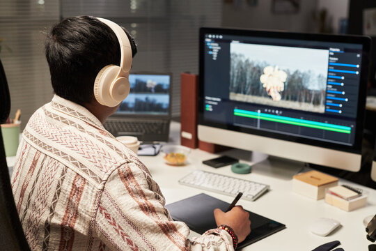 Young man editing video content on desktop computer while using pen tablet and headphones in home office. Useful for remote work, digital creator, postproduction marketing