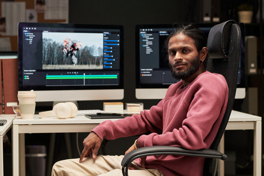 Portrait of young Indian man sitting at video editing workstation, reviewing footage on dual monitors in office. Useful for creative industry, post production, digital media marketing
