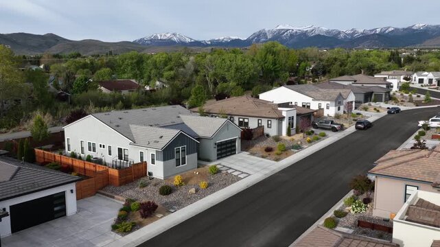 Modern desert-style homes line quiet street in Reno, Nevada, with xeriscaped front yards and snow-capped Sierra Nevada mountains rising above tree-filled valley behind. Aerial drone flyover shot