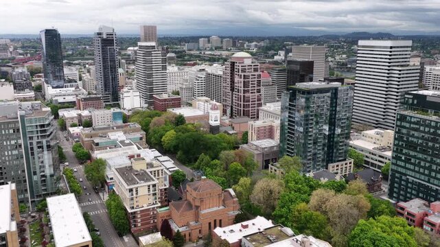 Downtown high-rise glass towers and historic church rise above tree-lined streets in Portland, Oregon, with rooftop pool and parked cars beneath cloudy hillside skyline. Aerial drone flyover shot