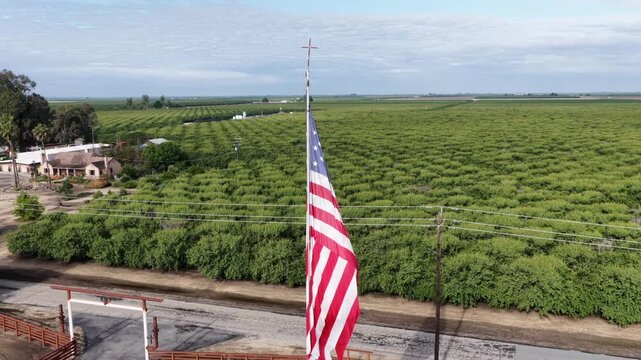 American flag stands beside orchard rows and white ranch house near Chowchilla, California, framed by dirt road, gate entrance and eucalyptus trees under overcast sky. Aerial drone wide shot