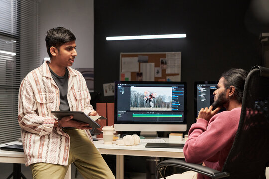 Two multiethnic men discussing video editing project at computer workstation, reviewing creative workflow and teamwork. Ideal for media production, agency collaboration, postproduction