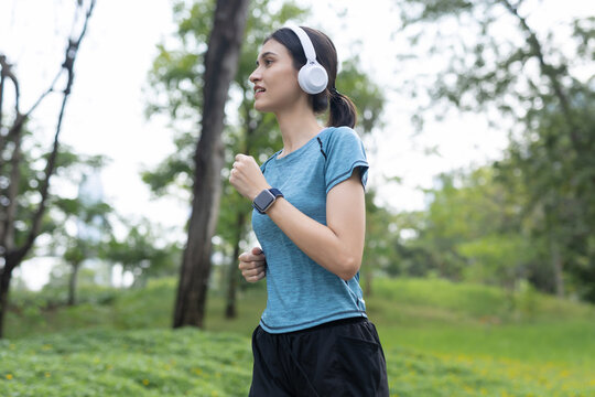 fit woman running in green public park with white headphone and modern smartwatch feeling happy and focused during morning fitness workout routine surrounded by beautiful nature environment