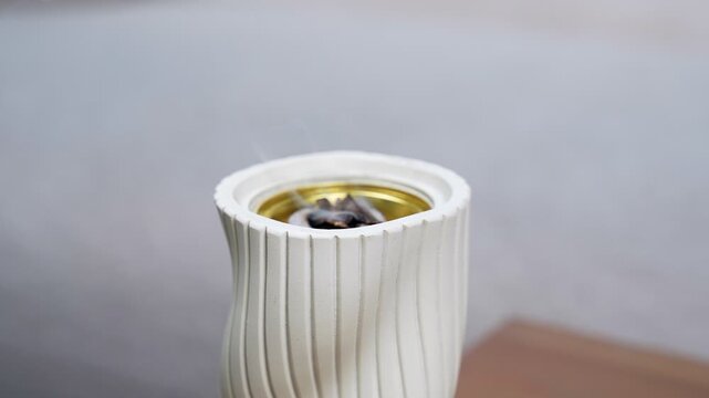 Close-up of burning oud wood chips inside an elegant white ceramic incense burner. Delicate white smoke rises from the gold interior against a blurred indoor background during natural daylight.