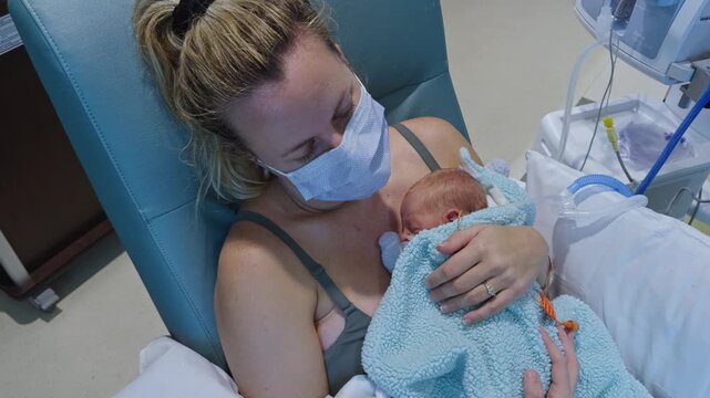 Masked mother holding a premature newborn baby skin to skin in kangaroo care position in a NICU hospital room.