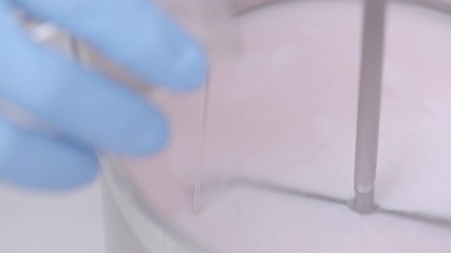Close-up of a hand with a blue protective glove pouring a liquid mixture of pale pink hue and uniform texture into a container, while stirring with a rod