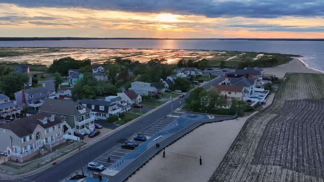 Aerial view of a coastal neighborhood in Union Beach, New Jersey at sunset with houses, a beach volleyball court, parking lot, and calm water beneath dramatic clouds.